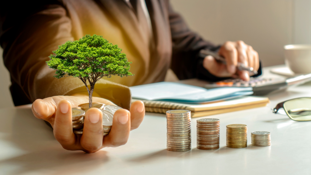 a person who is making notes is sitting with stacks of coins on front of them. their right hand is extended and is holding more coins with a tree coming out of them symbolizing a result of future investments