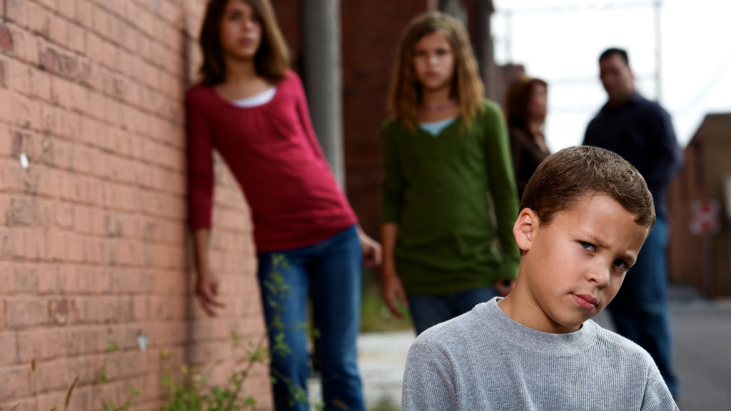 a group of three children, all looking towards the camera. the boy in the the front plan and in the background are two girls and what seems to be their parents in the third plan