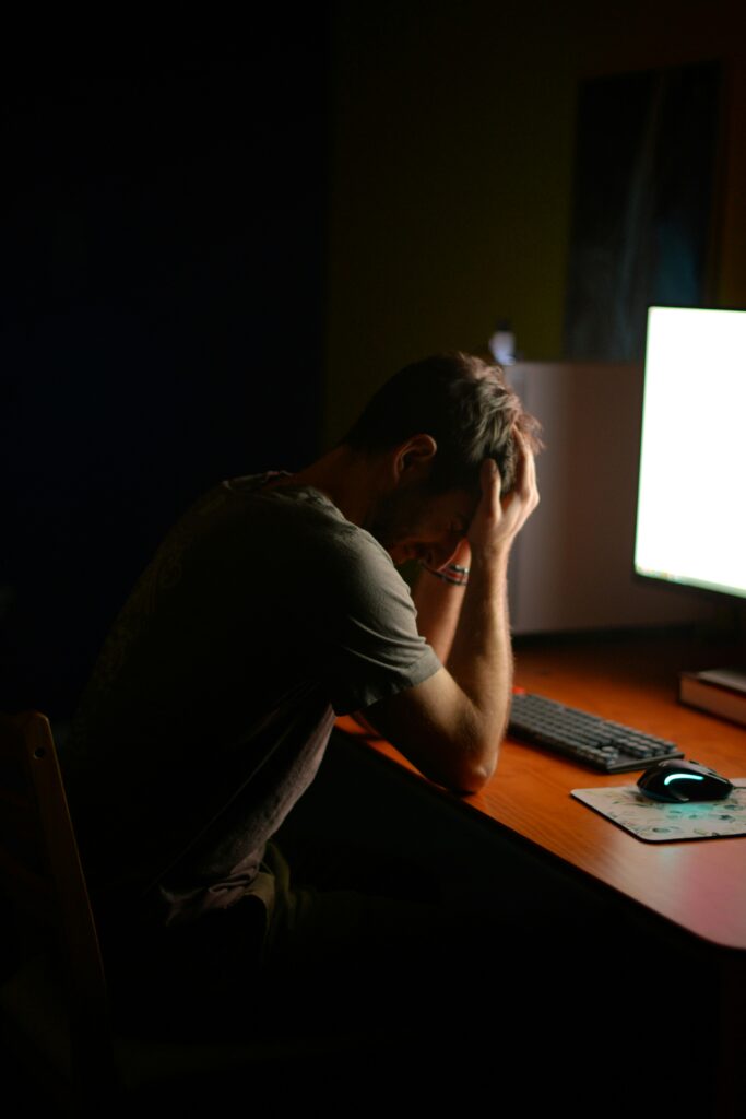 A man sitting at a desk in front of his computer with his head in his hands as if being tired, frustrated or with a headache