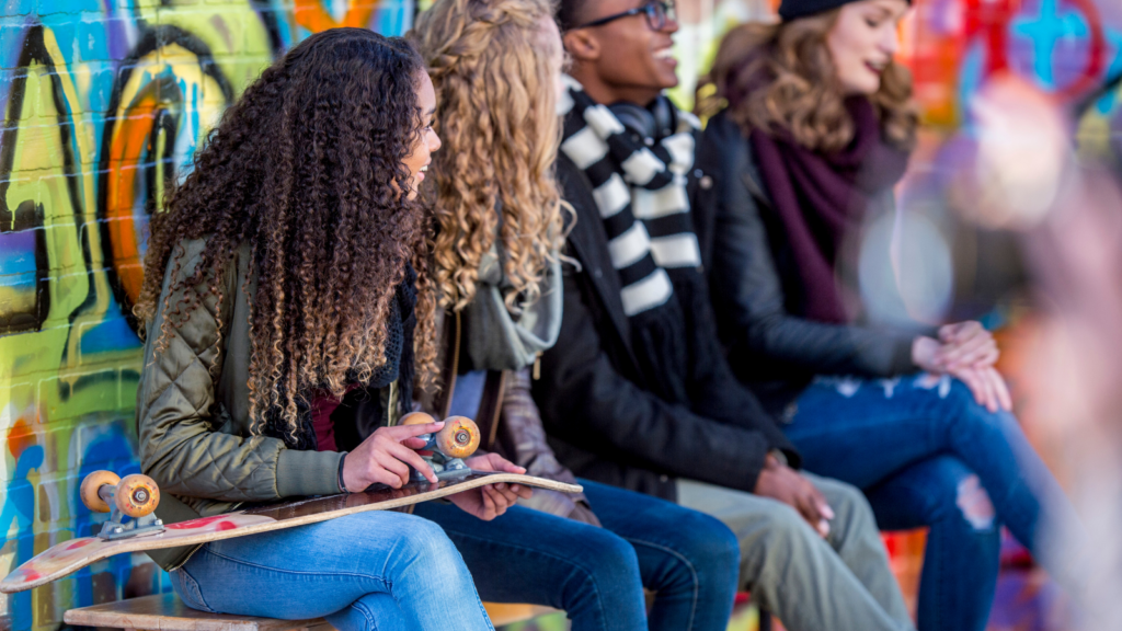 four teenagers; three girls and boy sit at an outdoor skatepark