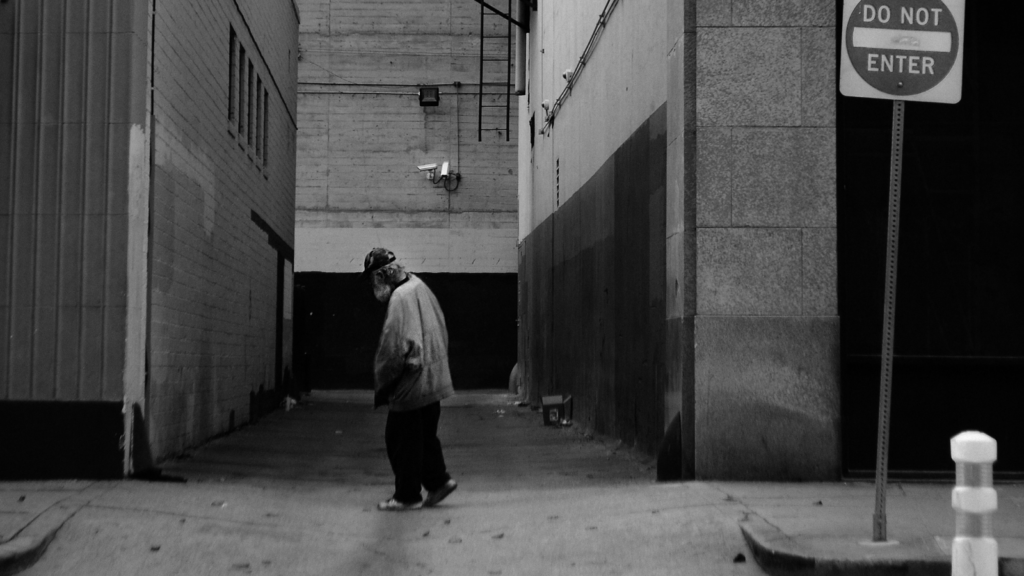 black and white: an elderly man who is possibly a rough sleeper walks the street while a sign reads "do not enter"