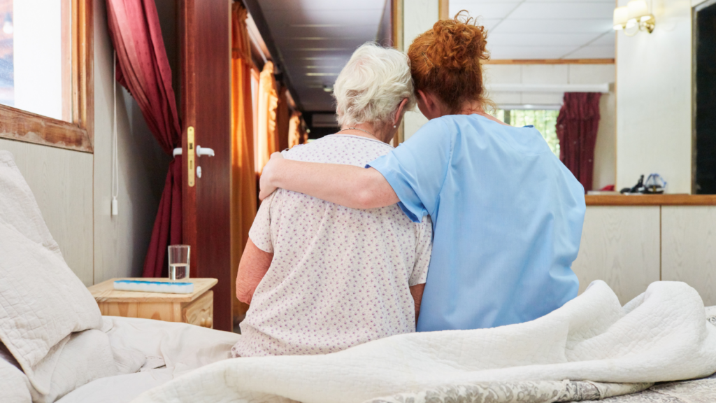 A female care worker puts her arm around an elderly patient as both sit on the edge of a bed in a care facility